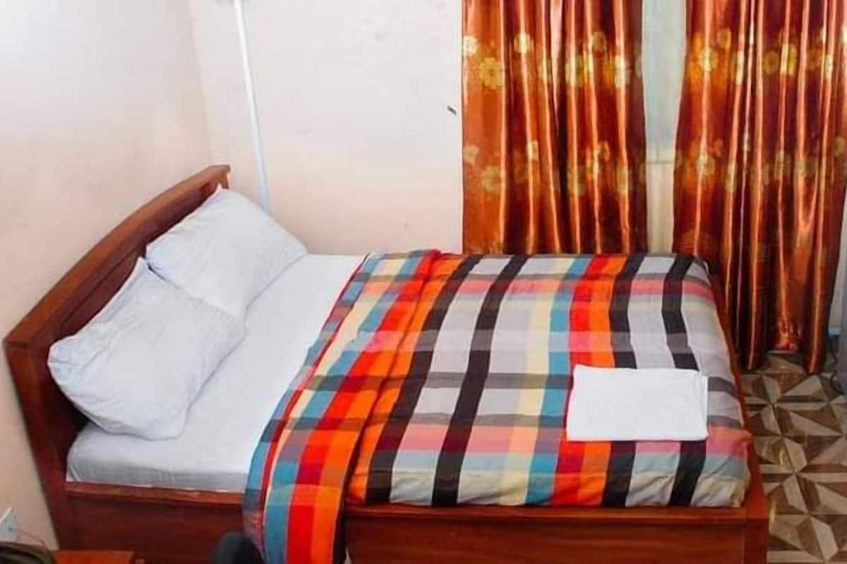 A single bed with a plaid bedspread, a white pillow, and a white blanket. There is a wooden headboard and a small table beside the bed. The room has a tiled floor and a curtain with a floral pattern.