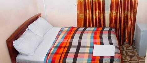 Un lit simple avec une couvre-lit à carreaux, un oreiller blanc et une couverture blanche. Il y a une tête de lit en bois et une petite table à côté du lit. La pièce a un sol carrelé et un rideau à motifs floraux.