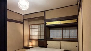 A traditional Japanese-style room with tatami flooring, a low bed with a white mattress, a small bedside table with a lamp, and shoji screens on the windows. - Guesthouse Hayashiya (Omachi)