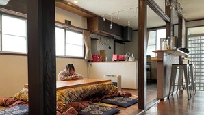 A cozy room with a wooden floor, a seating area with cushions, a kitchen area with a sink and cabinets, and a dining area with stools. - Guesthouse Hayashiya (Omachi)