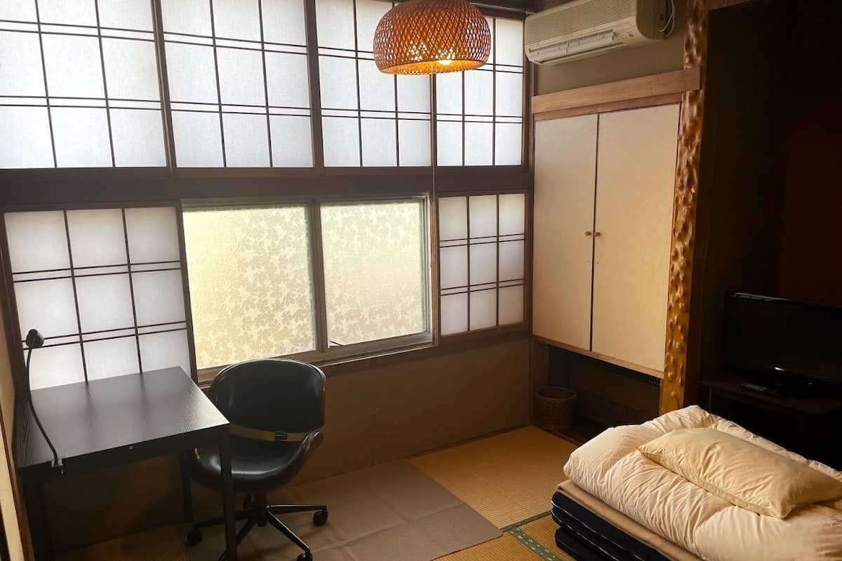 A room with tatami mat flooring, a desk with a chair, a bed with pillows, a television, and a traditional Japanese-style window with a hanging lamp.