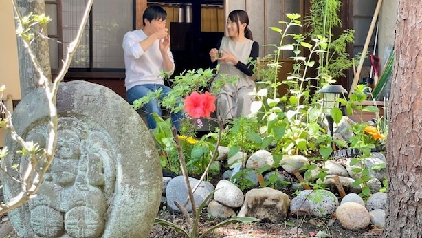 A traditional Japanese garden with a stone statue, a red flower, and a wooden building in the background. - Guesthouse Hayashiya (Omachi)