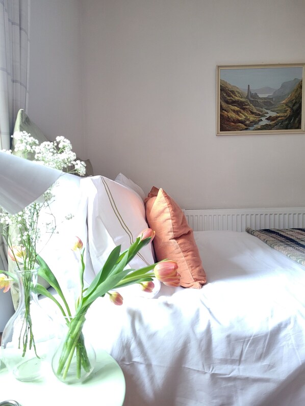 Una cama bien hecha con ropa de cama blanca y una almohada rosa dorado, un florero con flores sobre una mesita de noche y un cuadro en la pared.
