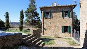 A stone building with two windows and a chimney, surrounded by trees and a stone wall. - Villa Mille Primavere (Gaiole in Chianti)
