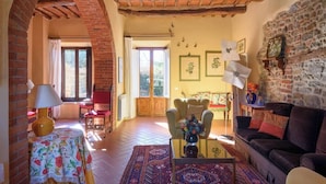 A cozy living room with a brick fireplace, a sofa, a coffee table with a vase, and a dining area with a table and chairs. - Villa Mille Primavere (Gaiole in Chianti)