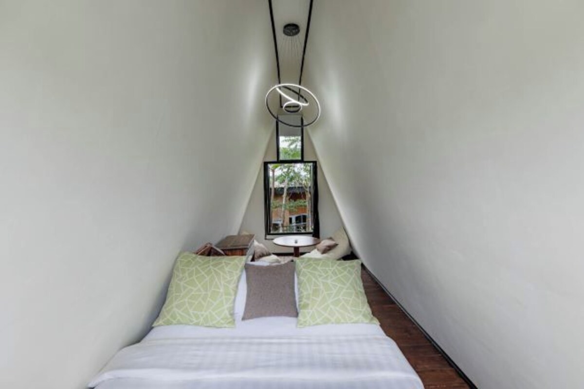 Una habitación estrecha y triangular con una cama al fondo, una pequeña mesa con una silla, y una ventana con vistas a árboles.