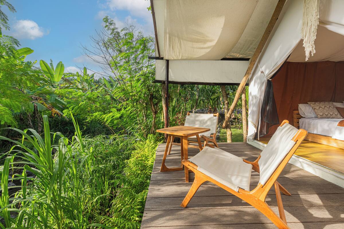 An outdoor wooden deck with a bed and two chairs, surrounded by lush greenery.