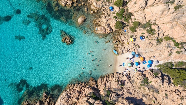 An aerial view of a rocky coastal area with clear turquoise water, a sandy beach, and scattered rocks.