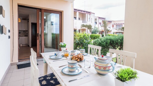 A dining table set with plates, cups, and a teapot on a balcony with a view of neighboring buildings and greenery. - Residence I Mirti n. 8 (Santa Teresa di Gallura)