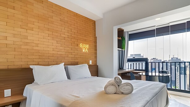 Een moderne hotelkamer met een geaccentueerde bakstenen wand, een groot bed met witte lakens, een netjes opgemaakt bed met opgerolde handdoeken, een kleine tafel met stoelen, en een balkon met uitzicht over de stad.
