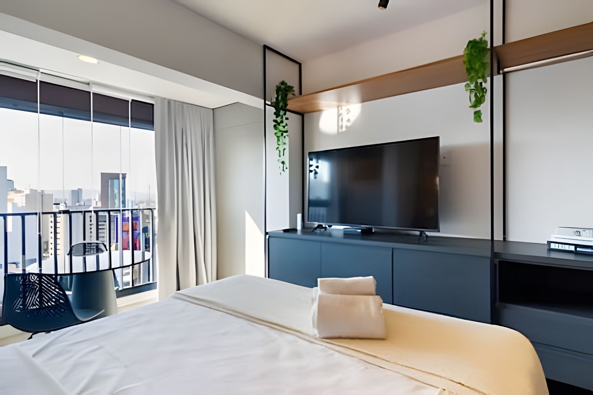 Une chambre d’hôtel moderne dotée d’un grand lit, d’un téléviseur à écran plat fixé au mur, d’un balcon offrant une vue sur la ville et d’un bureau avec un ordinateur.