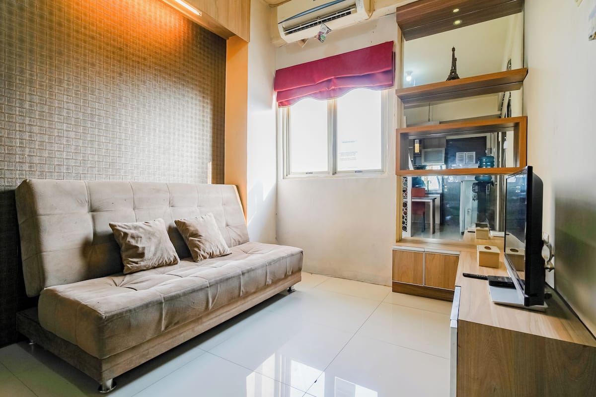 Una sala de estar moderna con un sofá color crema, un televisor de pantalla plana y una cocina visible a través de un separador de vidrio.