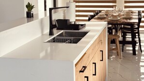 Une cuisine moderne avec un plan de travail blanc, un robinet noir et des armoires en bois. On aperçoit à l’arrière un coin repas avec une table et des chaises.