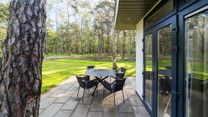 A patio with a round table and chairs, surrounded by a stone floor, overlooking a grassy area with trees. - Vacation Home in Haarle near Hellendoorn (Haarle)