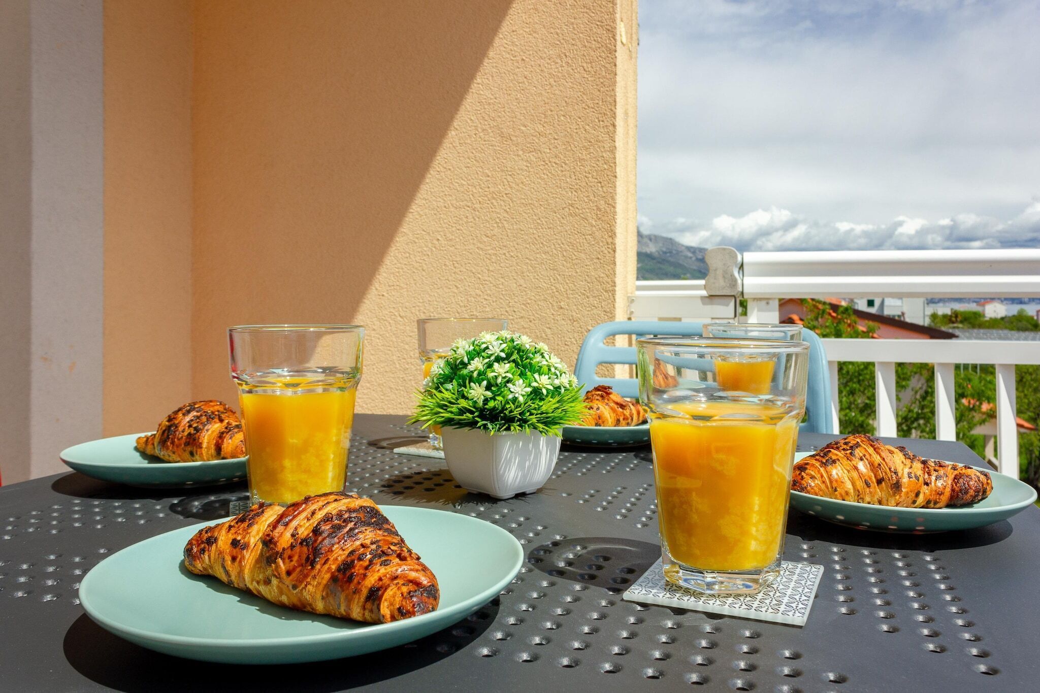 Une table avec des croissants, des verres de jus d’orange et une petite plante en pot, sur un balcon offrant une vue sur les montagnes.