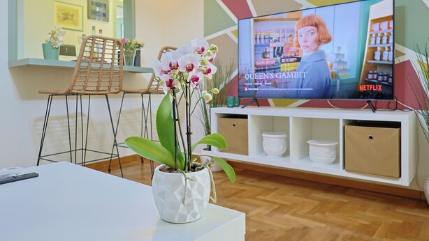 A living room with a TV displaying 'Queen's Gambit' on Netflix, a white sofa, a white coffee table, and a potted orchid. - Faliron Urban Retreat (Palaio Faliro)