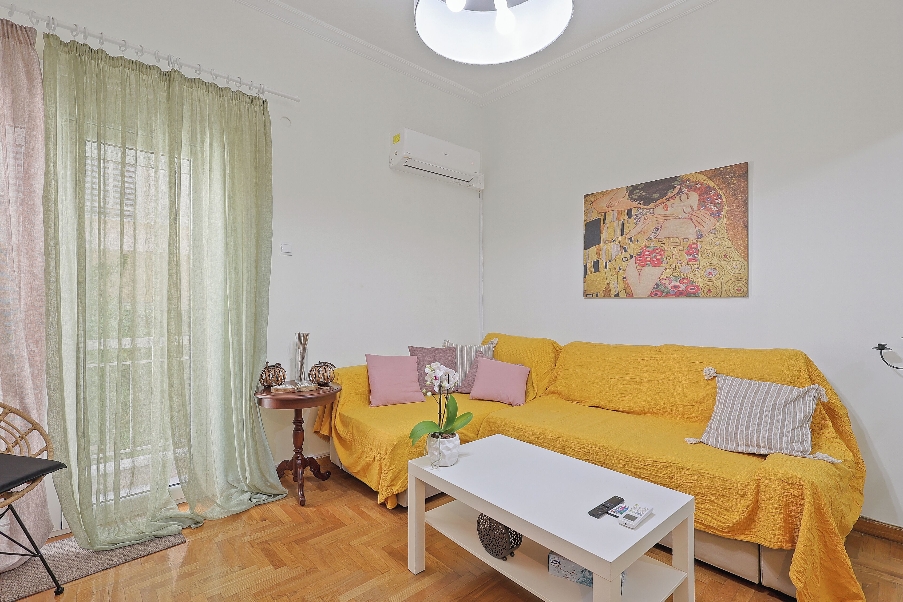A room with a yellow sofa, a white table, a small side table, a chair, and a painting on the wall.