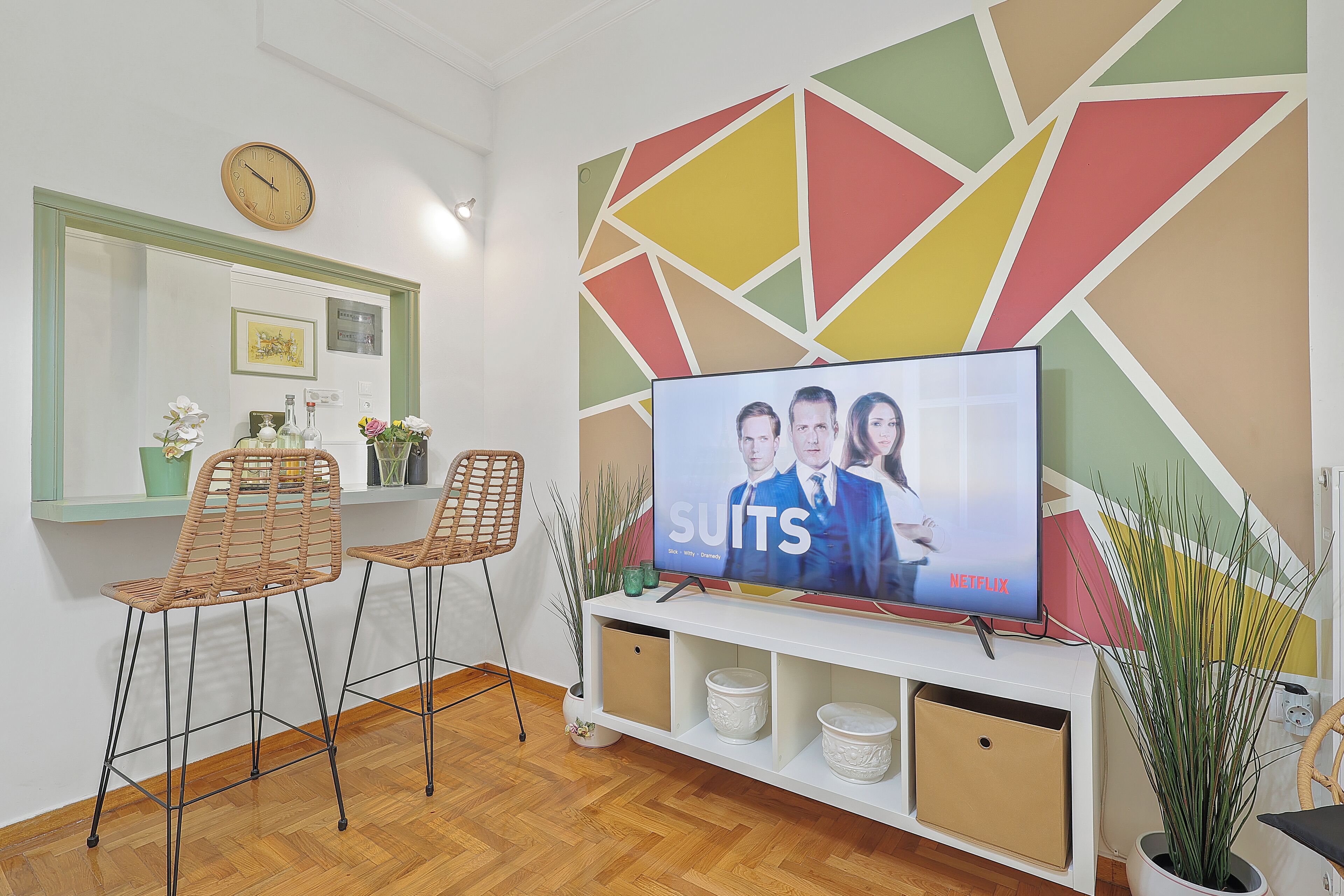 Un salon moderne doté d’un grand téléviseur à écran plat diffusant une série Netflix, de deux fauteuils en osier, d’une petite table agrémentée d’objets décoratifs et d’un motif géométrique sur le mur.