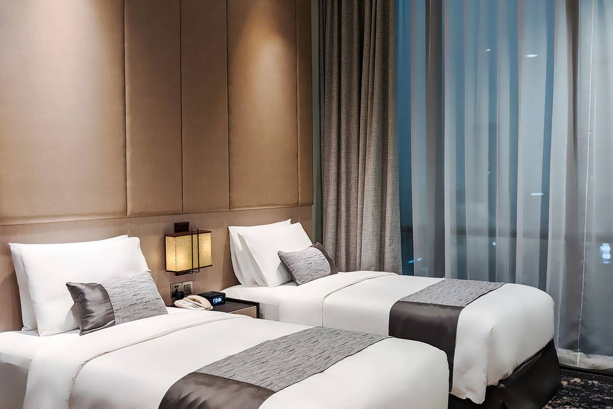 Habitación de hotel con dos camas, una mesita de noche con lámpara y vistas a la ciudad a través de cortinas amplias.