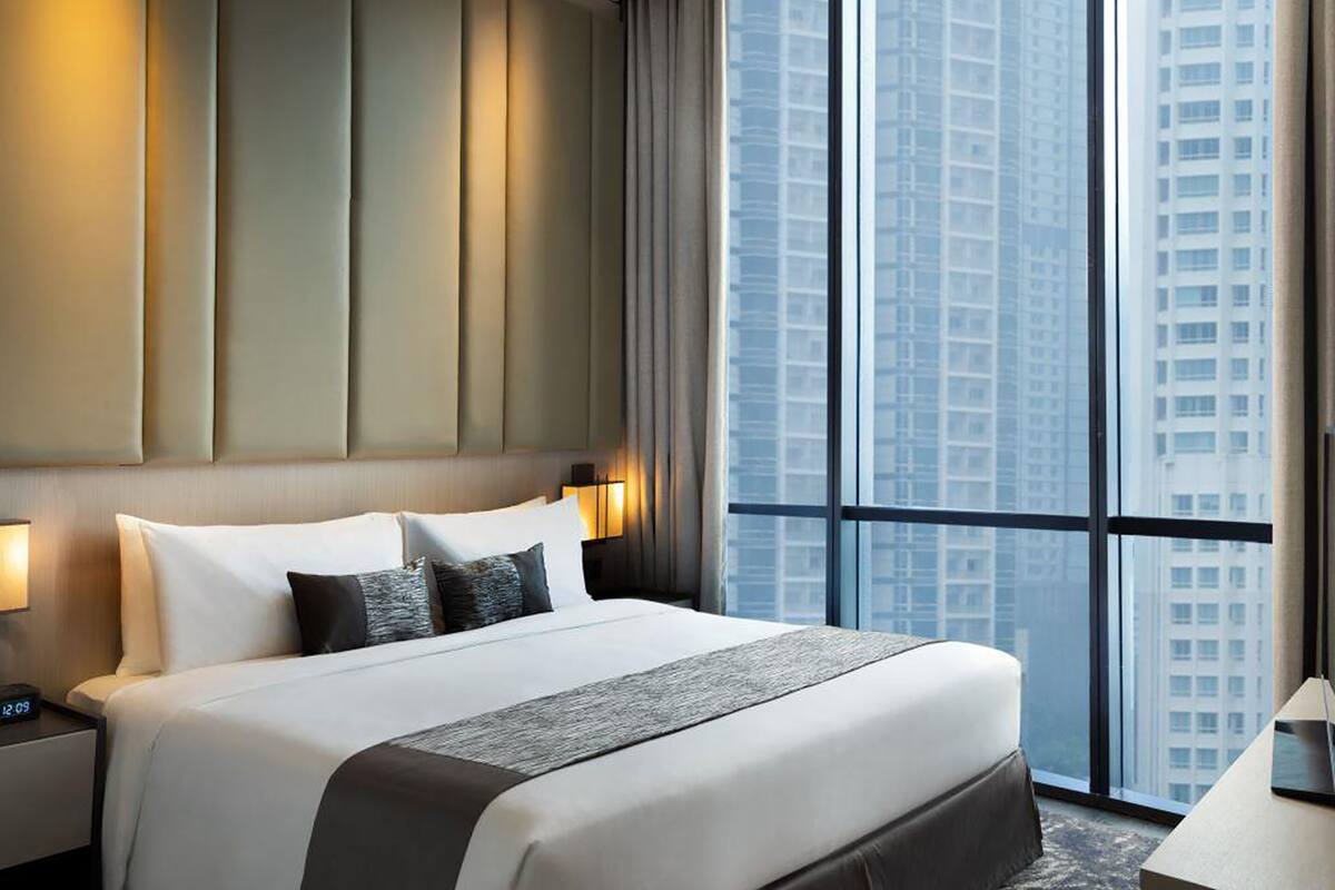 Una habitación de hotel moderna con una cama grande, una mesita de noche, una lámpara y vistas de edificios altos a través de amplias ventanas.