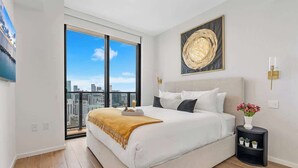 Quarto com uma cama grande, mesa de cabeceira, luminária na parede e vista da cidade através de uma janela ampla.