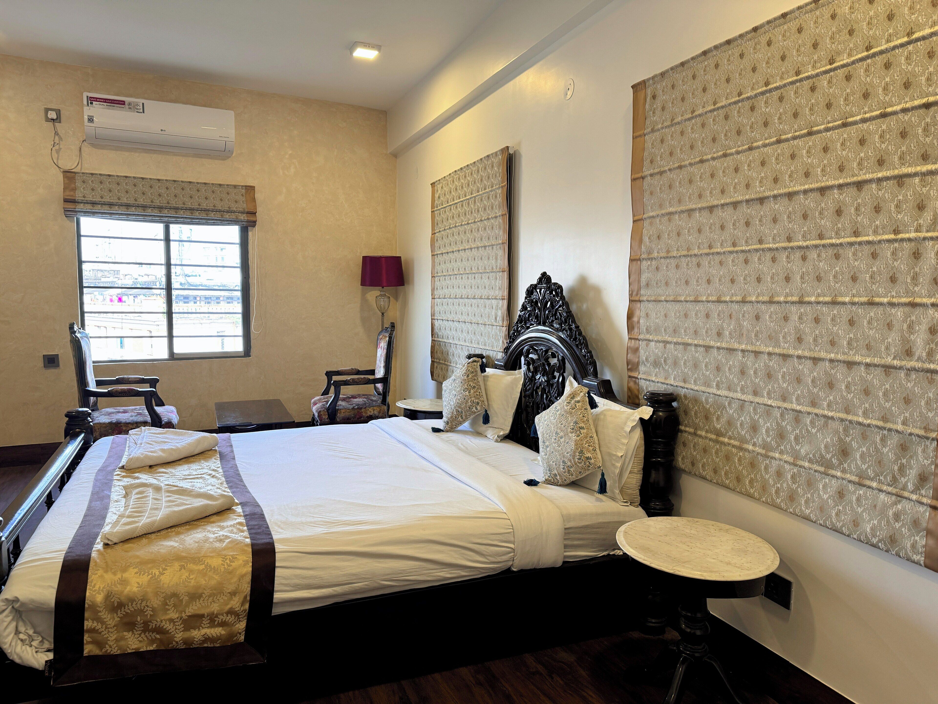 A hotel room with a large bed, a round table, a chair, an air conditioning unit, and a window with a view of buildings.