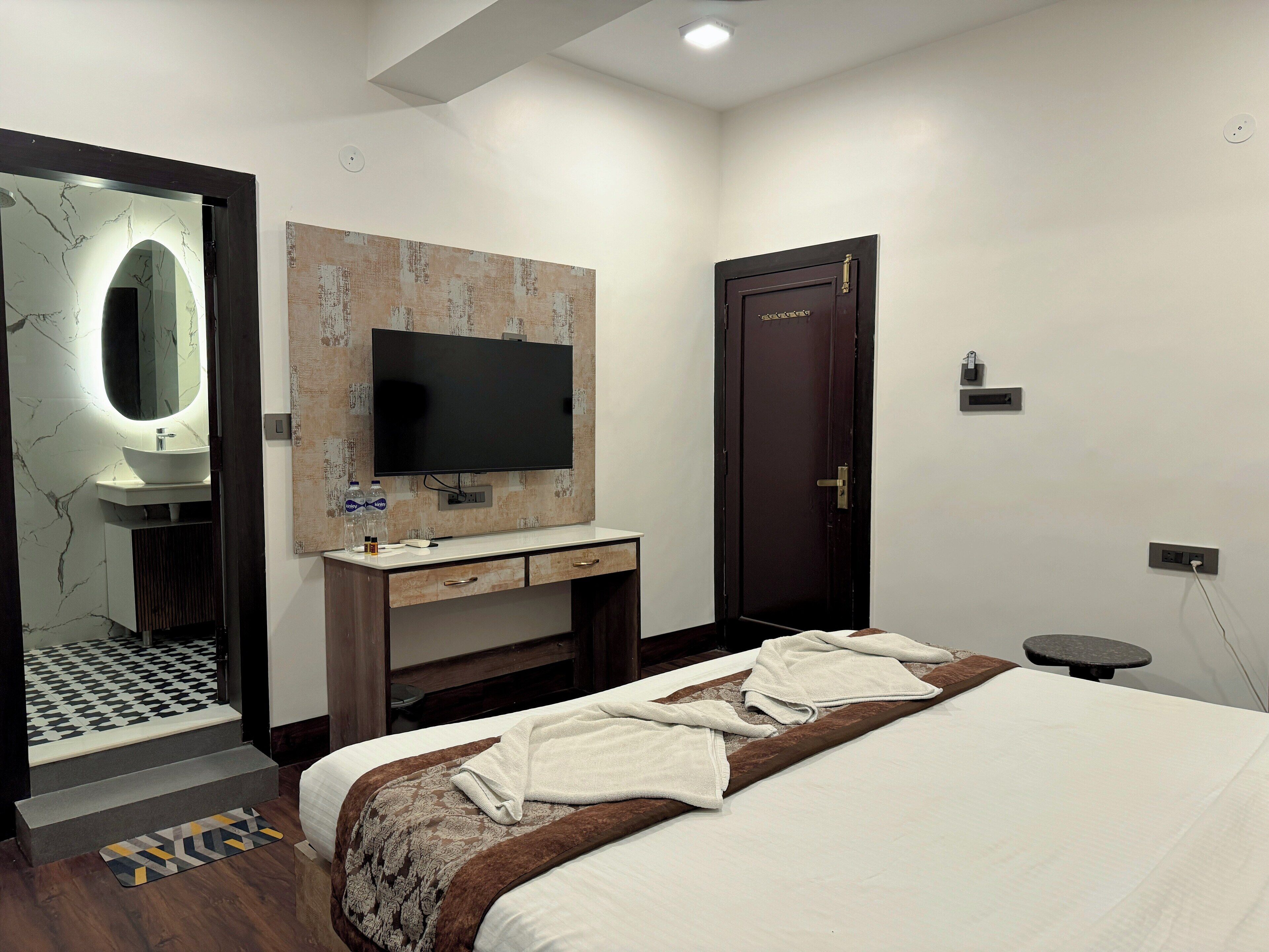 A modern hotel room with a large bed, a TV mounted on the wall, a wooden wardrobe, and a bathroom with a marble vanity.