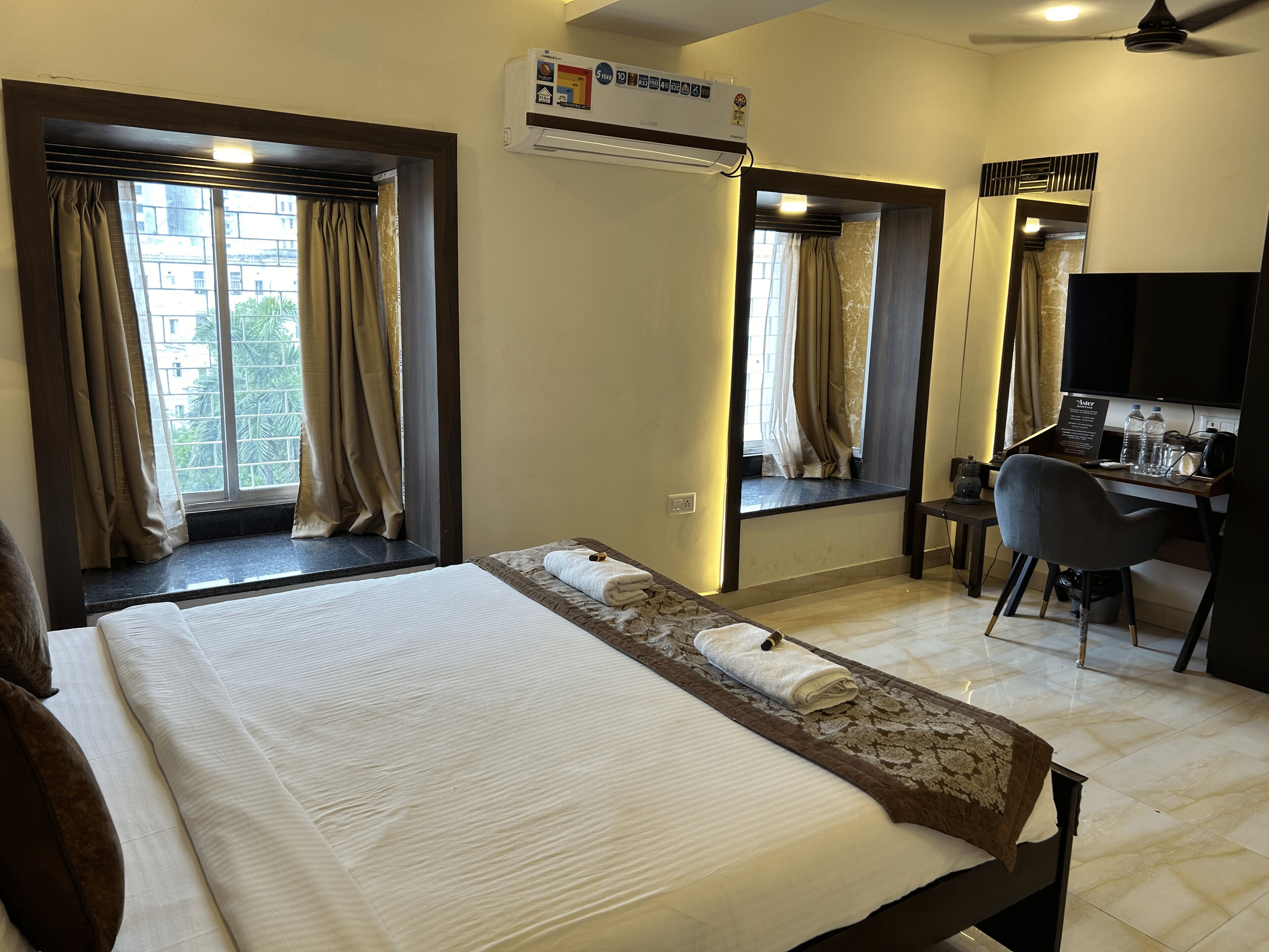 A hotel room with a large bed, a desk with a chair, a flat-screen TV, and an air conditioning unit.
