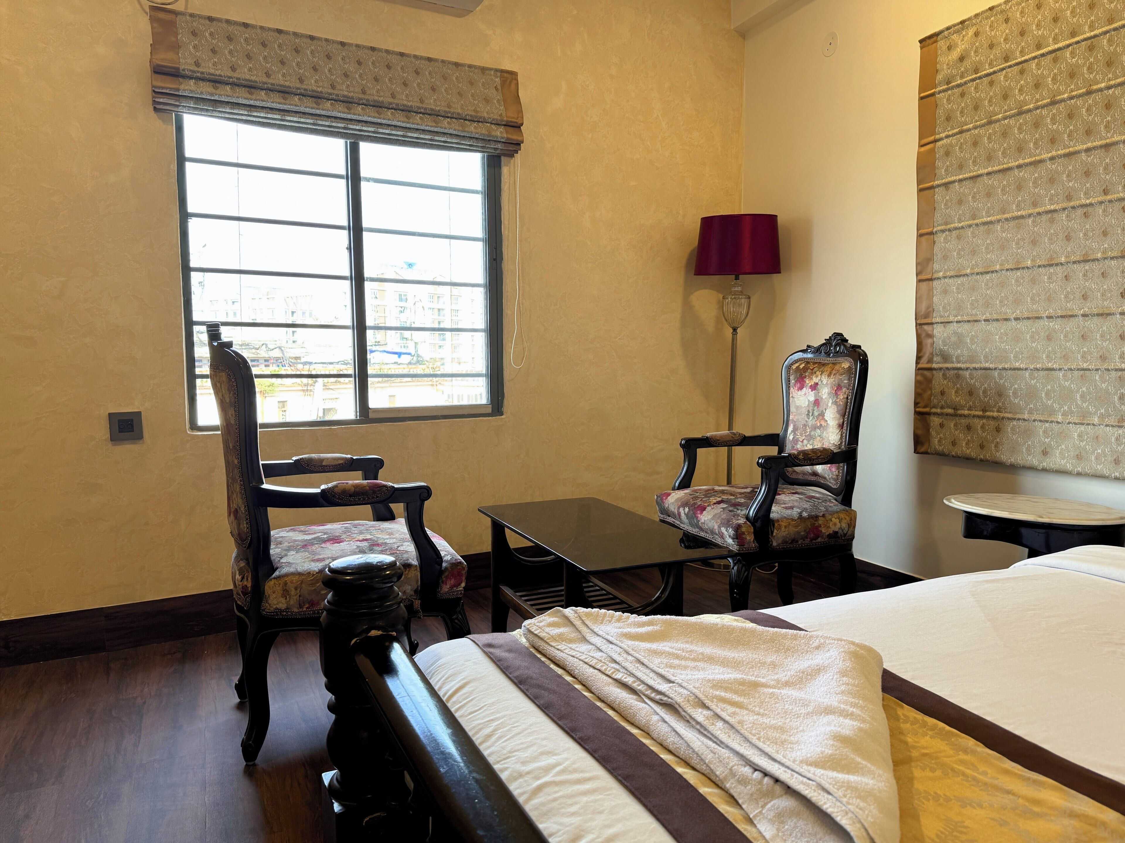 A hotel room with a bed, two chairs, a small table, a lamp, and a window with a view of buildings.