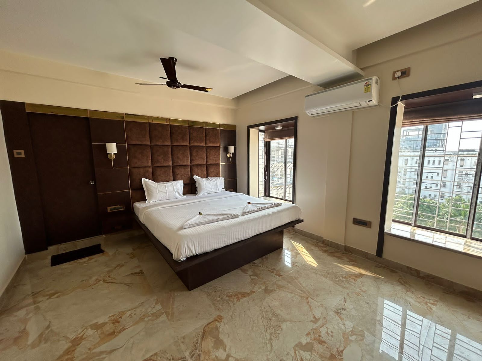 A modern bedroom with a large bed, a ceiling fan, an air conditioning unit, and a view of buildings through a window.