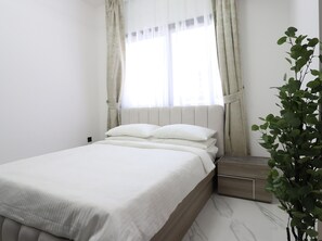Siististi pedattu sänky valkoisilla vuodevaatteilla huoneessa, jossa on ikkuna, yöpöytä ja ruukussa oleva kasvi.