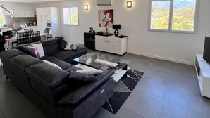 Un salón amplio con un sofá seccional, una mesa de centro de vidrio y una zona de comedor, con una cocina al fondo.