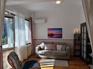 A living room with a sofa, a radiator, a window with curtains, a picture on the wall, and a lamp on a shelf.