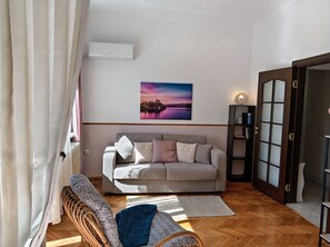 Una sala de estar con un sofá, una silla de mimbre, un radiador, un cuadro en la pared y una puerta con paneles de vidrio esmerilado.