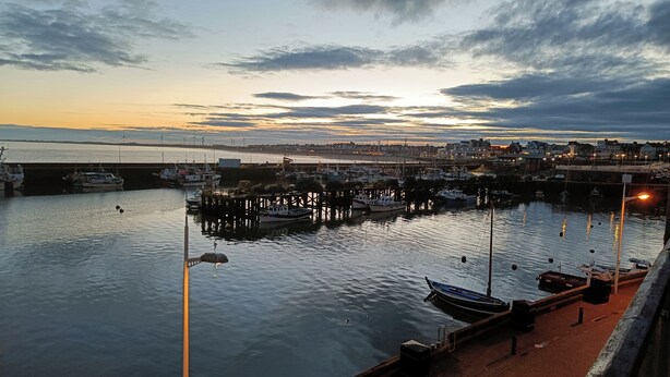 Un port où des bateaux sont amarrés aux quais, une surface d’eau calme et, au loin, une vue sur la ville sous un ciel nuageux.
