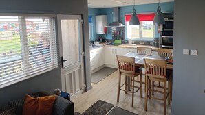 Une cuisine moderne avec des meubles blancs, un coin repas avec des chaises en bois et une grande fenêtre équipée de stores.