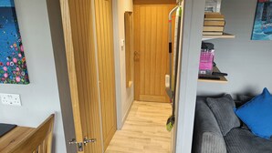 Un couloir avec des portes en bois, un balai appuyé contre un mur, un canapé avec un coussin bleu et une étagère avec des livres et un tableau.
