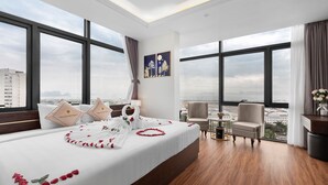 A hotel room with a large bed, two pillows, a nightstand, a chair, a small table, and a view of the city. - Thai Ha Boutique Hotel (Bai Chay)
