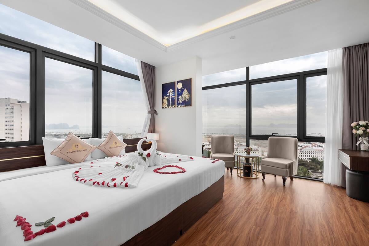 A hotel room with a large bed, two pillows, a nightstand, a chair, a small table, and a view of the city.