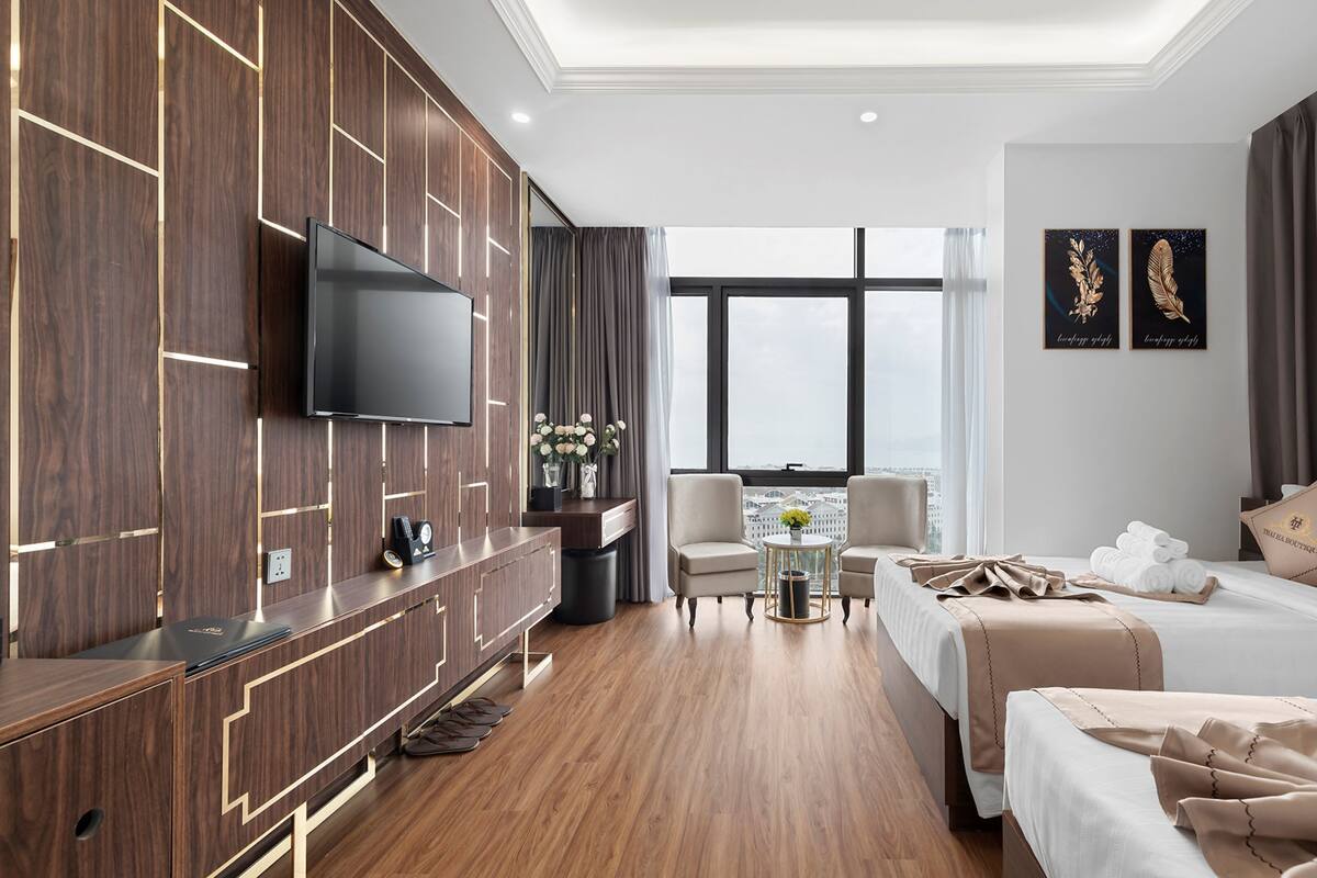 A modern hotel room with a large TV, a desk with a computer, two chairs, a bed, and a view of the city.