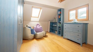 A room with a sloped ceiling, wooden beams, a skylight, a blue dresser, a bookshelf, and a window.