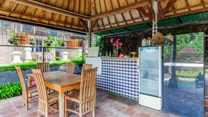 A covered outdoor dining area with a wooden table and chairs, a refrigerator, and a checkered tablecloth. - Belvilla Ratna Resort Ubud PalaceNearUbud Palace (Ubud)