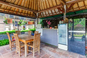 A covered outdoor dining area with a wooden table and chairs, a refrigerator, and a checkered tablecloth. - Belvilla Ratna Resort Ubud PalaceNearUbud Palace (Ubud)