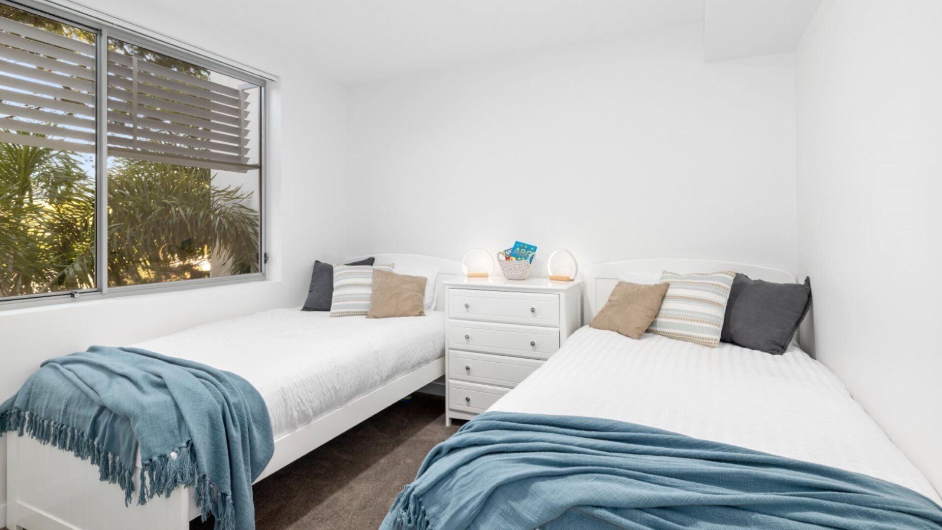 Un dormitorio con dos camas individuales, una mesita de noche con lámpara y una ventana con persianas.