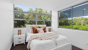A bedroom with a large bed, white nightstands, and a view of greenery through the windows. - Sounds of the Ocean on Boyd (WOORIM)