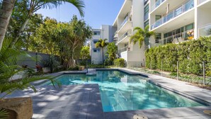 A modern pool area with a clear blue pool, surrounded by lush greenery and a multi-story building with balconies. - Sounds of the Ocean on Boyd (WOORIM)