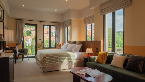 Un cuarto de hotel con una cama grande, un sofá verde, un escritorio y vistas a edificios a través de las ventanas.