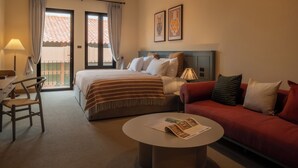 Habitación de hotel con cama, sofá rojo, mesa de centro redonda, escritorio con silla y balcón con cortinas.