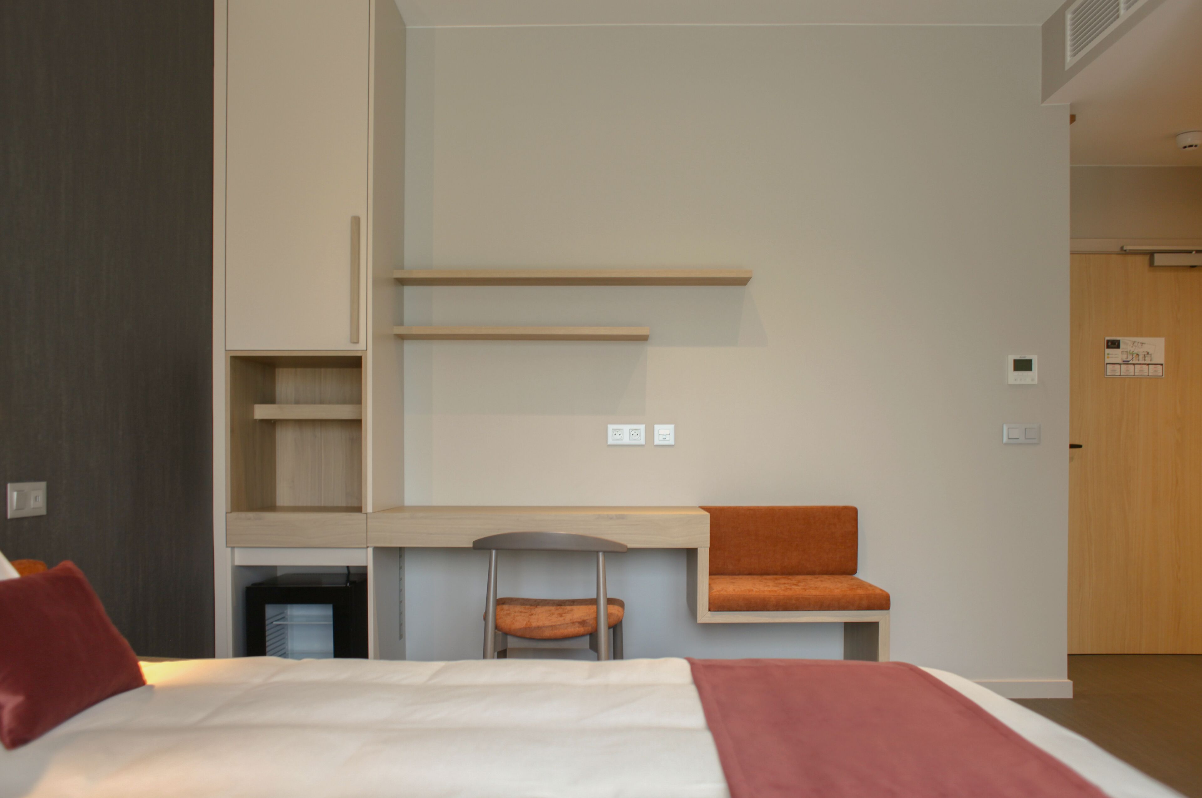 A modern hotel room with a bed, a desk, a chair, and a small table with a cushion.