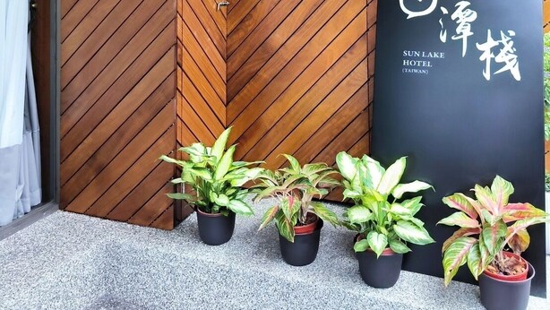 A wooden partition with a chevron pattern, a black sign with white text for Sun Lake Hotel in Taiwan, and several potted plants in front. - Sun lake Hotel (Yuchi)
